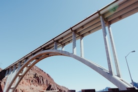 A large, modern concrete bridge with an arched support structure spans across a rocky landscape. The sky is clear and blue, providing bright and natural lighting to the scene. The architectural design of the bridge emphasizes its strength and elegance.
