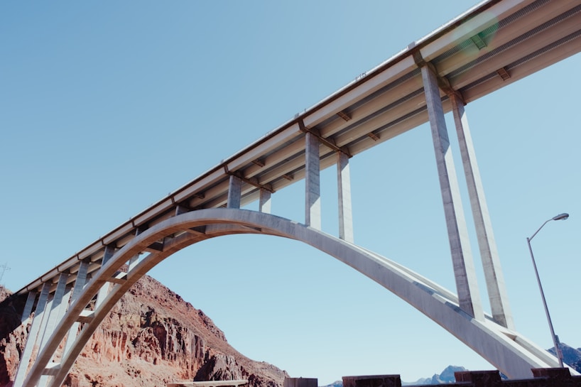A large, modern concrete bridge with an arched support structure spans across a rocky landscape. The sky is clear and blue, providing bright and natural lighting to the scene. The architectural design of the bridge emphasizes its strength and elegance.