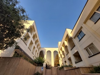 A modern, symmetrical building with large arches and white walls, framed by a clear blue sky. The building is flanked by trees, and a sign indicating accessible parking is visible in the foreground.