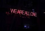 A neon sign with glowing red letters spells out 'WE ARE ALL ONE' against a dark background. The sign is mounted on what appears to be a building structure, partially obscured by shadows and reflections.