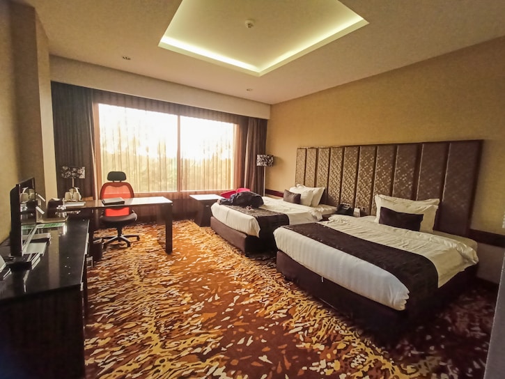 A hotel room featuring two neatly made single beds with dark brown and white bedding. The room has a unique patterned carpet with shades of brown and beige. A desk is placed near the large window, accompanied by an orange and black office chair. The window allows natural light to brighten the room, and the curtains are partially drawn. There is a sleek, dark-toned furniture setup, including a cabinet and a small kitchen area with a coffee maker.