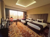 A hotel room featuring two neatly made single beds with dark brown and white bedding. The room has a unique patterned carpet with shades of brown and beige. A desk is placed near the large window, accompanied by an orange and black office chair. The window allows natural light to brighten the room, and the curtains are partially drawn. There is a sleek, dark-toned furniture setup, including a cabinet and a small kitchen area with a coffee maker.