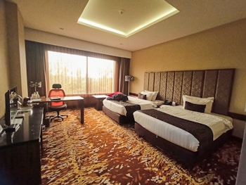 A hotel room featuring two neatly made single beds with dark brown and white bedding. The room has a unique patterned carpet with shades of brown and beige. A desk is placed near the large window, accompanied by an orange and black office chair. The window allows natural light to brighten the room, and the curtains are partially drawn. There is a sleek, dark-toned furniture setup, including a cabinet and a small kitchen area with a coffee maker.