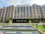 A modern, multi-story building with a symmetrical facade features large glass windows and beige exterior walls. In front, there are neatly trimmed shrubs and well-maintained landscaping. The entrance prominently displays the words 'THE PALACE' in bold letters, with large decorative planters on either side.