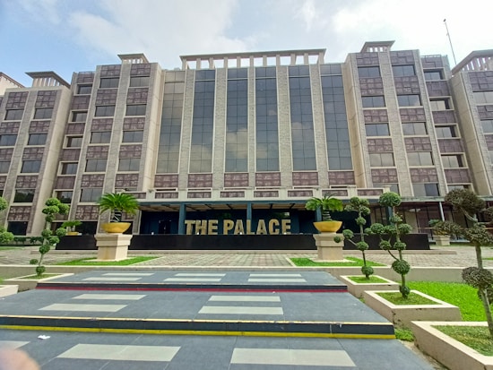 A modern, multi-story building with a symmetrical facade features large glass windows and beige exterior walls. In front, there are neatly trimmed shrubs and well-maintained landscaping. The entrance prominently displays the words 'THE PALACE' in bold letters, with large decorative planters on either side.