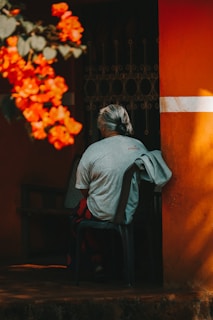 An elderly person with gray hair is sitting on a plastic chair near an orange wall. Bright red flowers are in the foreground, partially out of focus, adding contrast to the scene. The person, dressed in a light gray T-shirt with another shirt draped over the chair back, is viewed from behind, creating a sense of solitude.