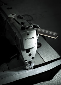A close-up view of a heavy-duty industrial sewing machine labeled 'TYPICAL'. The machine's needle and presser foot are shown engaging with dark fabric. The image features a metallic handle and various components that are typical in industrial sewing equipment.