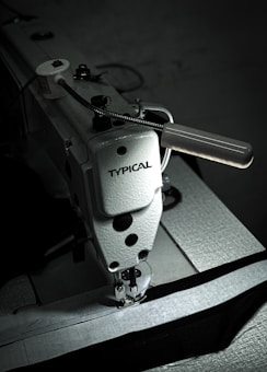A close-up view of a heavy-duty industrial sewing machine labeled 'TYPICAL'. The machine's needle and presser foot are shown engaging with dark fabric. The image features a metallic handle and various components that are typical in industrial sewing equipment.