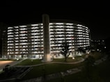 A large multi-story residential building with a curved facade is illuminated against the night sky. Each level is brightened by room lights, creating a grid-like pattern. In the foreground, well-maintained lawns with a few trees and a pathway lit by street lamps add to the serene nighttime setting.