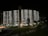 A large multi-story residential building with a curved facade is illuminated against the night sky. Each level is brightened by room lights, creating a grid-like pattern. In the foreground, well-maintained lawns with a few trees and a pathway lit by street lamps add to the serene nighttime setting.