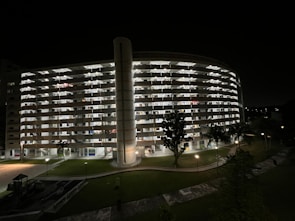 A large multi-story residential building with a curved facade is illuminated against the night sky. Each level is brightened by room lights, creating a grid-like pattern. In the foreground, well-maintained lawns with a few trees and a pathway lit by street lamps add to the serene nighttime setting.