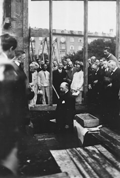A group of men is gathered around a construction or renovation site, with one man kneeling down in the center. The scene appears formal, likely a ceremony or official event, with several men in suits and a few in work attire. The background includes scaffolding and a partially constructed or damaged building facade, with visible greenery and an adjacent building.
