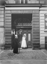 Two women stand in front of an ornate, arch-topped wooden doorway set into a stone building facade. One woman wears a dark outfit with a headdress and holds a walking stick, while the other is dressed in a white uniform, possibly a nurse, also with a white headdress. They appear to be engaged in conversation, standing on a cobblestone street. The building features detailed masonry and large rectangular windows.