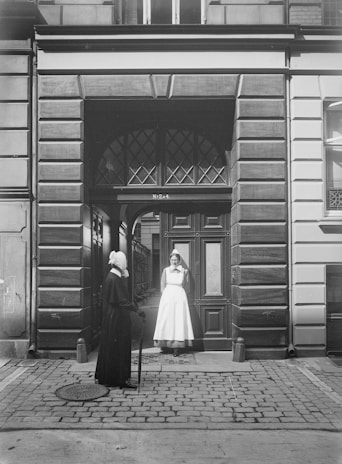 Two women stand in front of an ornate, arch-topped wooden doorway set into a stone building facade. One woman wears a dark outfit with a headdress and holds a walking stick, while the other is dressed in a white uniform, possibly a nurse, also with a white headdress. They appear to be engaged in conversation, standing on a cobblestone street. The building features detailed masonry and large rectangular windows.