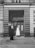 Two women stand in front of an ornate, arch-topped wooden doorway set into a stone building facade. One woman wears a dark outfit with a headdress and holds a walking stick, while the other is dressed in a white uniform, possibly a nurse, also with a white headdress. They appear to be engaged in conversation, standing on a cobblestone street. The building features detailed masonry and large rectangular windows.