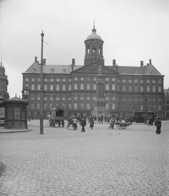 The image depicts a historical city square with cobblestone pavement, surrounded by a grand classical building with a clock tower and numerous windows. Several people are walking and interacting in the open space, and there are horse-drawn carriages present.
