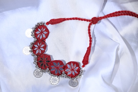 A handcrafted necklace lies on a white fabric background. The necklace features an intricate design with red, star-shaped patterns connected by a braided red thread. Small silver-colored coins and decorative beads are attached along the edge, creating a striking contrast against the red elements.