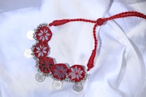 A handcrafted necklace lies on a white fabric background. The necklace features an intricate design with red, star-shaped patterns connected by a braided red thread. Small silver-colored coins and decorative beads are attached along the edge, creating a striking contrast against the red elements.