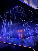 An illuminated stage featuring vertical strands of LED lights, emitting blue and purple hues, is the centerpiece. The geometric patterns on the floor reflect the light, enhancing the futuristic ambiance. In the background, a structure displays glowing text, adding depth to the scene.
