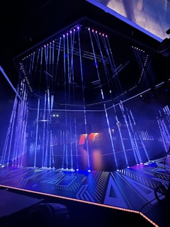 An illuminated stage featuring vertical strands of LED lights, emitting blue and purple hues, is the centerpiece. The geometric patterns on the floor reflect the light, enhancing the futuristic ambiance. In the background, a structure displays glowing text, adding depth to the scene.