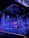 An illuminated stage featuring vertical strands of LED lights, emitting blue and purple hues, is the centerpiece. The geometric patterns on the floor reflect the light, enhancing the futuristic ambiance. In the background, a structure displays glowing text, adding depth to the scene.