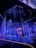 An illuminated stage featuring vertical strands of LED lights, emitting blue and purple hues, is the centerpiece. The geometric patterns on the floor reflect the light, enhancing the futuristic ambiance. In the background, a structure displays glowing text, adding depth to the scene.