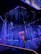 An illuminated stage featuring vertical strands of LED lights, emitting blue and purple hues, is the centerpiece. The geometric patterns on the floor reflect the light, enhancing the futuristic ambiance. In the background, a structure displays glowing text, adding depth to the scene.