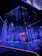 An illuminated stage featuring vertical strands of LED lights, emitting blue and purple hues, is the centerpiece. The geometric patterns on the floor reflect the light, enhancing the futuristic ambiance. In the background, a structure displays glowing text, adding depth to the scene.