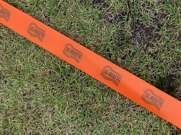 A vibrant orange tape stretches across a patch of green grass, featuring a repeating logo with stylized text. The tape contrasts sharply with the natural grass, which appears lush and slightly damp in areas.