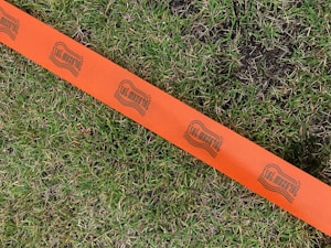 A vibrant orange tape stretches across a patch of green grass, featuring a repeating logo with stylized text. The tape contrasts sharply with the natural grass, which appears lush and slightly damp in areas.