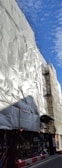 A building covered with white protective sheeting, surrounded by scaffolding. The blue sky with scattered clouds provides a contrasting background. There are images and advertisements covering the lower part of the structure.