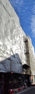 A building covered with white protective sheeting, surrounded by scaffolding. The blue sky with scattered clouds provides a contrasting background. There are images and advertisements covering the lower part of the structure.