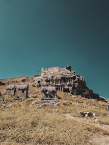 Ancient stone structures and tombs carved into rocky terrain, with a clear blue sky above. The scene captures the remnants of a historical site, surrounded by dry grass and scattered stones.