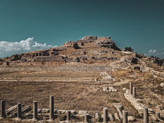 Ancient ruins sit atop a hill under a clear sky with scattered clouds. Stone structures and columns are partially buried among the dry grass, indicative of a historical archaeological site.