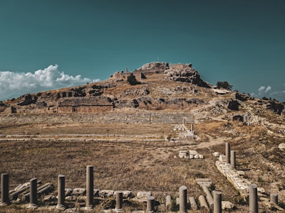 Ancient ruins sit atop a hill under a clear sky with scattered clouds. Stone structures and columns are partially buried among the dry grass, indicative of a historical archaeological site.