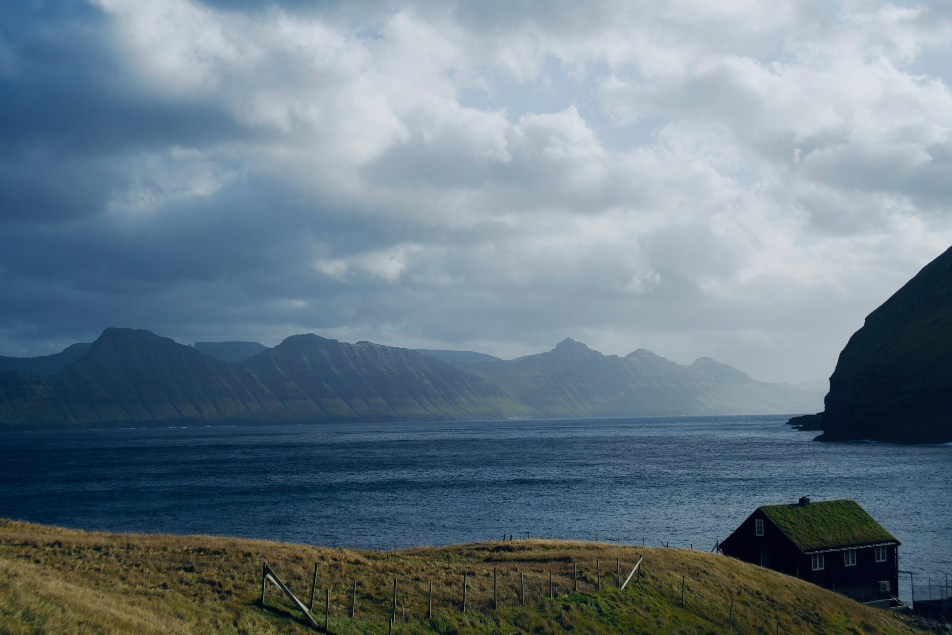 A serene landscape featuring a calm sea bordered by rugged, mountainous terrain in the background. A lone house with a grass-covered roof sits on a gentle slope of lush green grass, partially shadowed by surrounding hills. The sky is filled with thick, dramatic clouds, suggesting overcast weather with patches of sunlight breaking through.