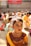 A young child dressed in traditional Indian attire, including a vibrant yellow and red dress adorned with jewelry and makeup. The background shows a group of children seated on the floor, also in traditional outfits. Banners in another language are visible on the walls.