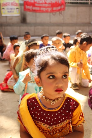 A young child dressed in traditional Indian attire, including a vibrant yellow and red dress adorned with jewelry and makeup. The background shows a group of children seated on the floor, also in traditional outfits. Banners in another language are visible on the walls.
