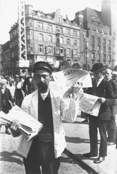 A street scene with a man wearing a cap and a white jacket holding a stack of newspapers in one hand and waving an open newspaper in the other. He is standing in front of a group of buildings with others dressed in period attire in the background.