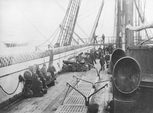 A black-and-white photograph depicts a busy ship deck populated by several crew members dressed in naval uniforms. The deck is lined with equipment, including ropes and cannons. The ship’s masts and rigging are visible, while crew members stand and attend to various tasks.