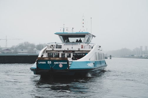A ferry with blue and white coloring travels on a body of water under grey, overcast skies. The ferry is labeled 'IJVEER 55 AMSTERDAM' and carries passengers in an enclosed cabin on the upper deck. In the distance, a crane and blurred cityscape can be seen.