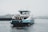 A ferry with blue and white coloring travels on a body of water under grey, overcast skies. The ferry is labeled 'IJVEER 55 AMSTERDAM' and carries passengers in an enclosed cabin on the upper deck. In the distance, a crane and blurred cityscape can be seen.