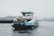 A ferry with blue and white coloring travels on a body of water under grey, overcast skies. The ferry is labeled 'IJVEER 55 AMSTERDAM' and carries passengers in an enclosed cabin on the upper deck. In the distance, a crane and blurred cityscape can be seen.