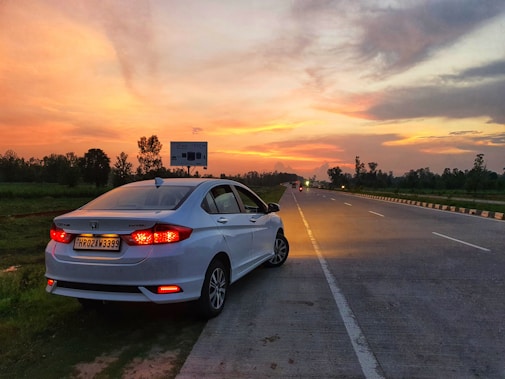 A silver sedan is parked on the side of a road during a vibrant sunset. The sky is filled with warm hues of orange, yellow, and pink, with scattered clouds. The road stretches into the distance, bordered by green grass and trees. The rear taillights of the car are illuminated.