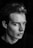 A black and white portrait of a young man with a serious expression on his face, gazing slightly away from the camera. His hair is styled back, and he wears a dark shirt, with dramatic lighting creating shadows that accentuate his facial features.
