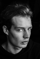 A black and white portrait of a young man with a serious expression on his face, gazing slightly away from the camera. His hair is styled back, and he wears a dark shirt, with dramatic lighting creating shadows that accentuate his facial features.