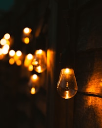 Warmly lit light bulbs are hanging against a dark wooden background, creating a cozy and inviting atmosphere. The soft glow of the bulbs contrasts with the shadows, highlighting the texture of the wood.