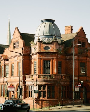 A historic red brick building with ornate architectural details, including a large, rounded dome on top, embellished with decorative elements. The structure appears to be aged, with some wear and vegetation growing along the edges. In front, there are signs indicating the building is for sale. A few cars are parked on the street in front of the building. A tall spire is visible in the background, suggesting an urban setting.