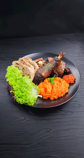 A black plate holds a serving of grilled meat garnished with herbs, accompanied by vibrant orange mashed carrots, fresh green lettuce, and slices of whole-grain bread. A small portion of bright orange fish roe adds a touch of elegance and color to the ensemble.
