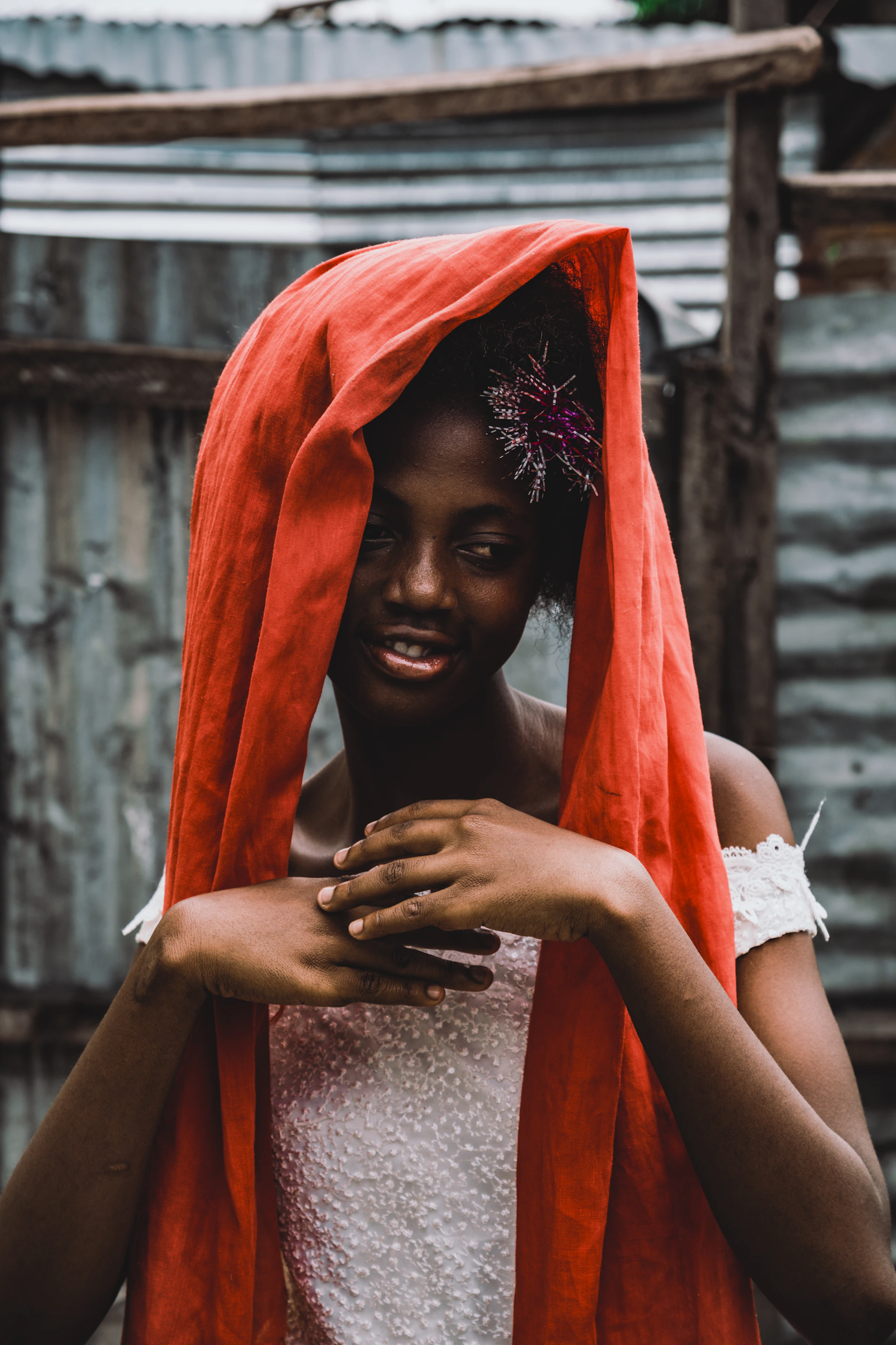 A person is wearing a red headscarf draped over their head, with a beaded or decorative piece near the left side of the face. Their hands are gracefully placed in front, and they are wearing an off-white textured garment. The background consists of corrugated metal, suggesting an outdoor setting.